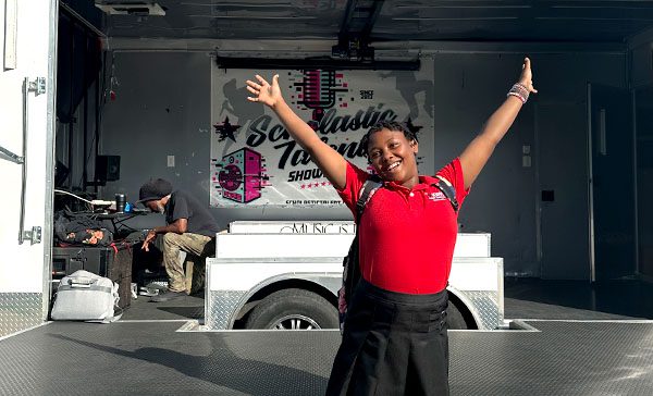 Young female scholar smiling and standing in front of mobile stage with hands in the air